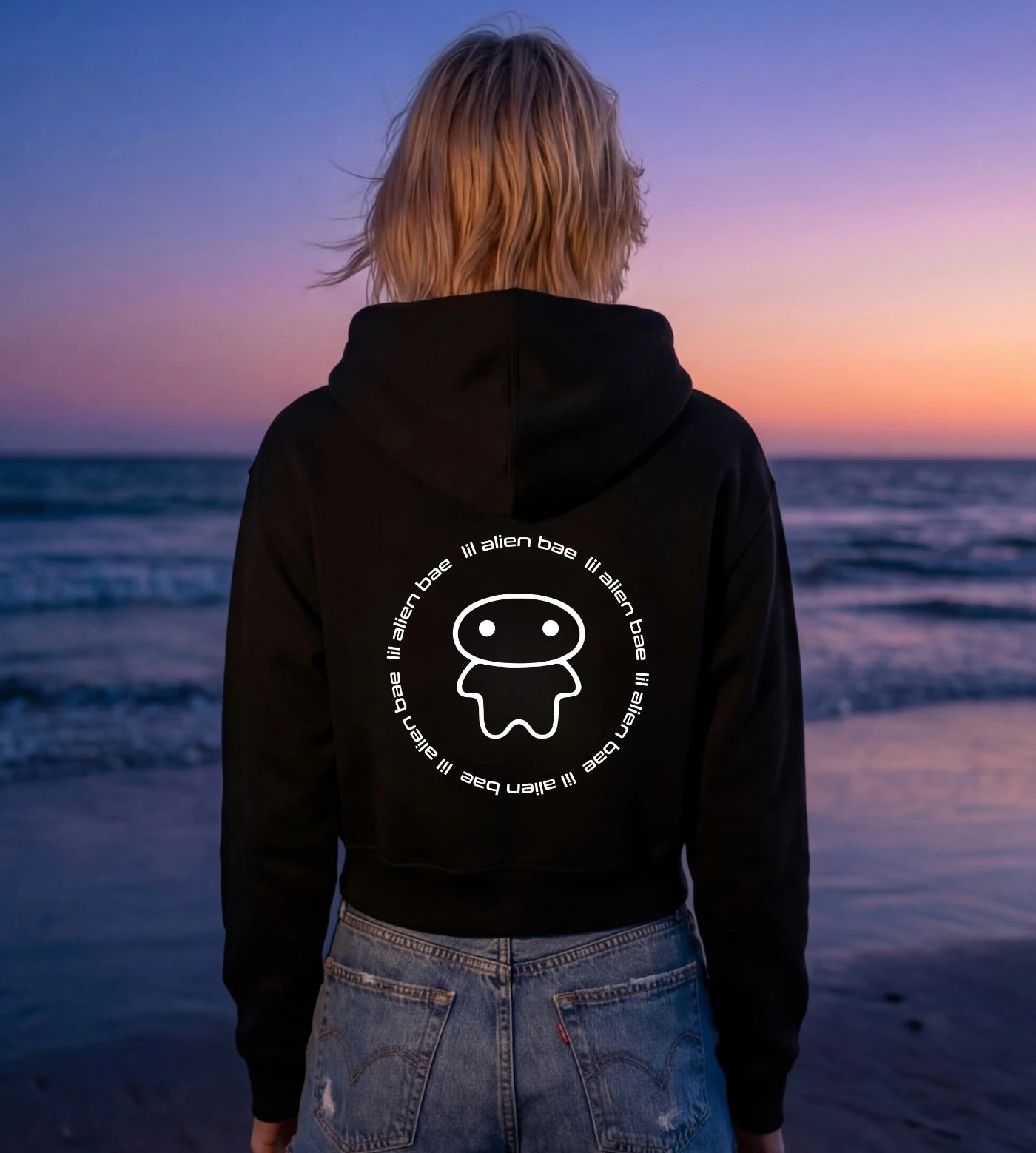 Model wears a black Cropped Fleece Zip-Up Hoodie featuring a Lil Alien Bae back graphic. 