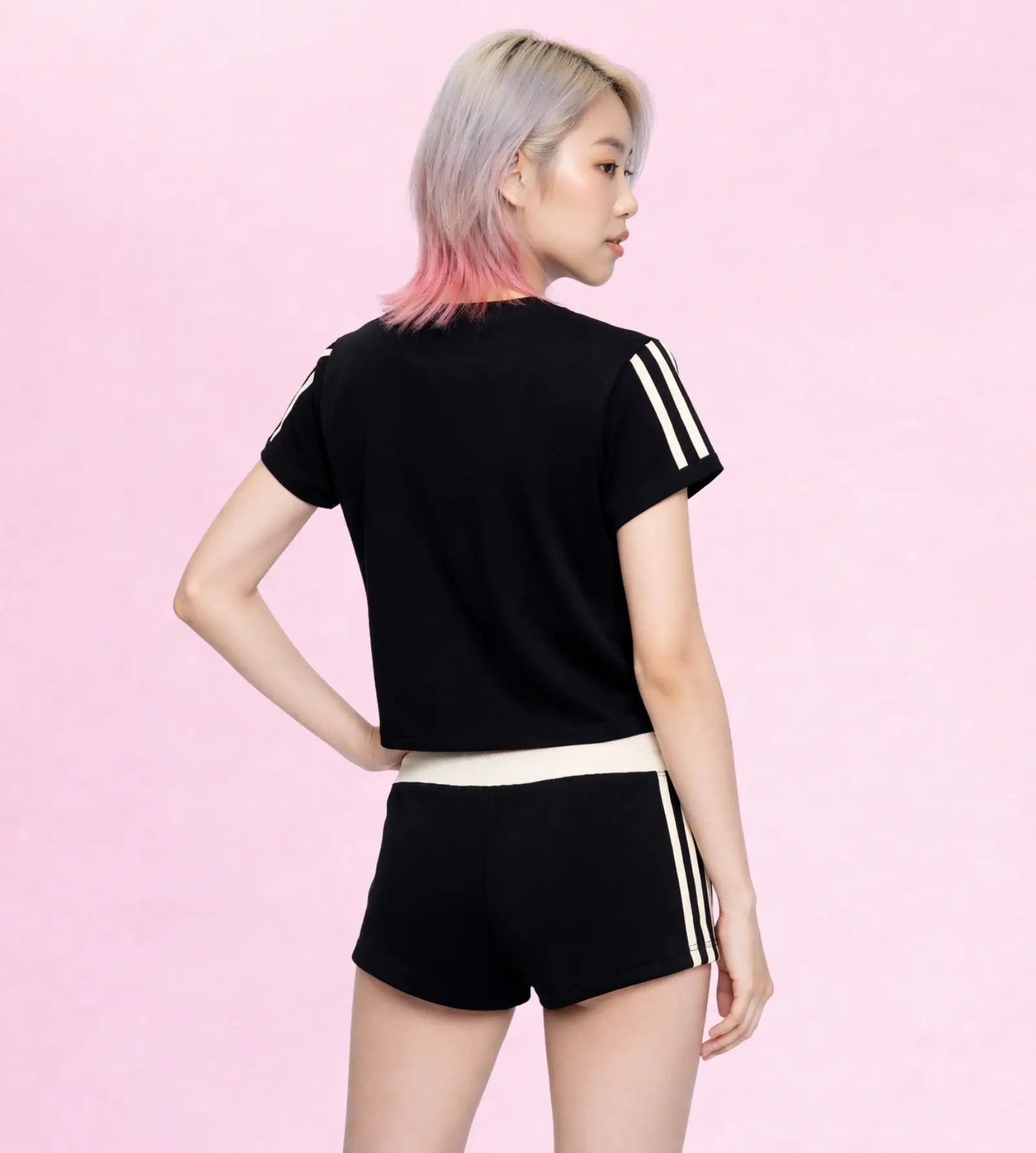 A model from the back wearing a black short-sleeve crop top and matching black athletic shorts, both featuring white three-stripe details on the sleeves and sides, set against a solid pink background.