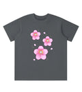 A dark gray short-sleeve t-shirt with a graphic print of three pink cherry blossom flowers and small pink petals on the front, displayed against a solid white background.