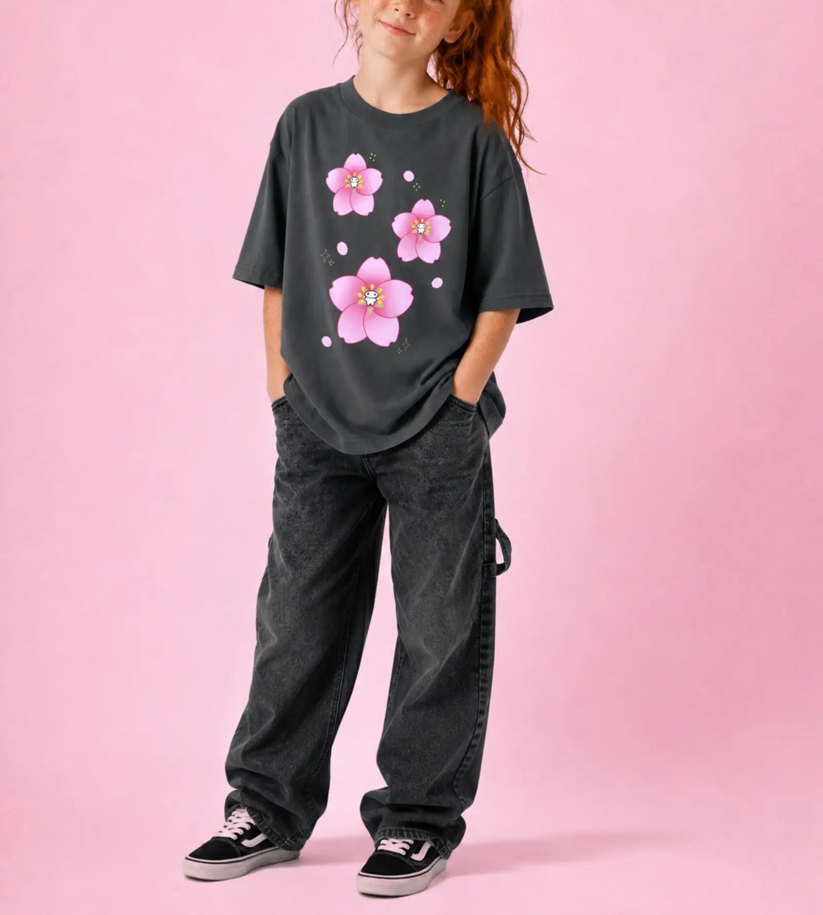 A child wearing a dark charcoal gray oversized t-shirt with a pink cherry blossom graphic, paired with dark gray washed jeans and black sneakers, standing against a solid pink background.