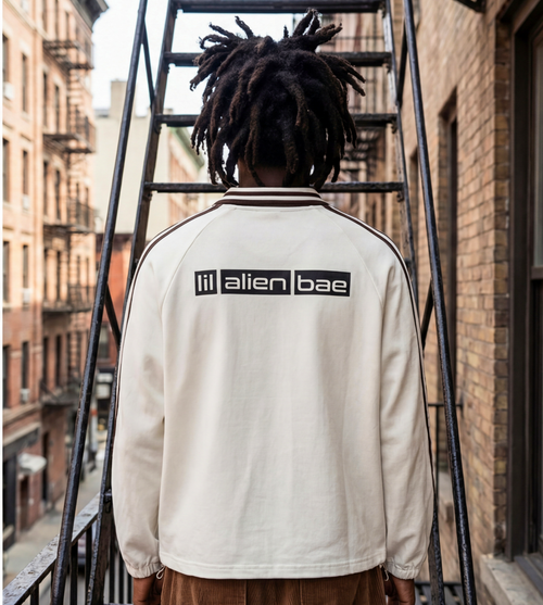 Model wearing an off white graphic pullover with a large lil alien bae logo across the back shoulders.  Outfit styled with brown corduroy pants.