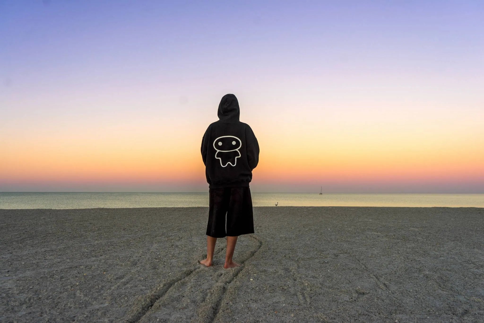 A person stands on a beach at sunrise facing the ocean, wearing a black hoodie with a lil alien bae graphic on the back and black shorts.