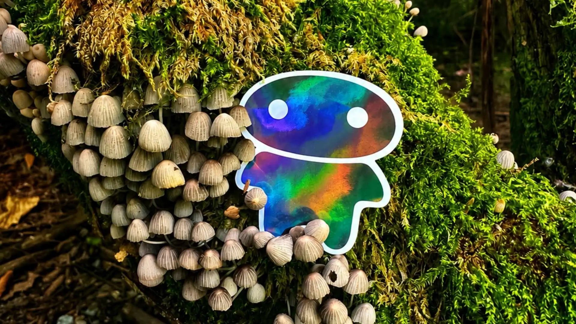 A die-cut sticker of a lil alien bae  with an iridescent rainbow finish and white outline, placed on a mossy log covered with small mushrooms in a forest setting.